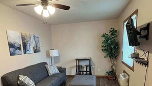 Un salon avec un ventilateur de plafond, un canapé gris, une petite table et une plante en pot.