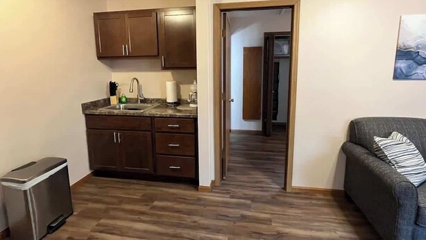 Un espace de vie compact comprenant un coin cuisine, un canapé gris et un plancher en bois.