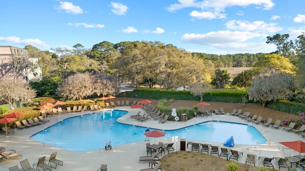 Relax by the serene pool surrounded by lush greenery and trees, with lounge chairs and umbrellas providing ample shade.