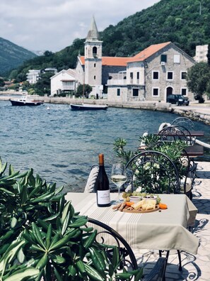 Uma mesa com uma garrafa de vinho, uma taça e um prato de queijo e uvas, à beira-mar, com barcos e uma igreja ao fundo.