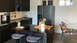 A modern kitchen with dark cabinets, a central island, bar stools, and a dining area with a glass table and metal chairs.