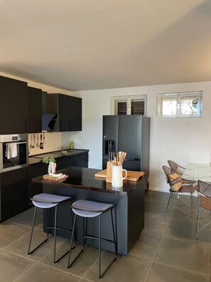 Una cocina moderna con armarios oscuros, una isla central, taburetes de bar y un comedor con mesa de vidrio y sillas de metal.