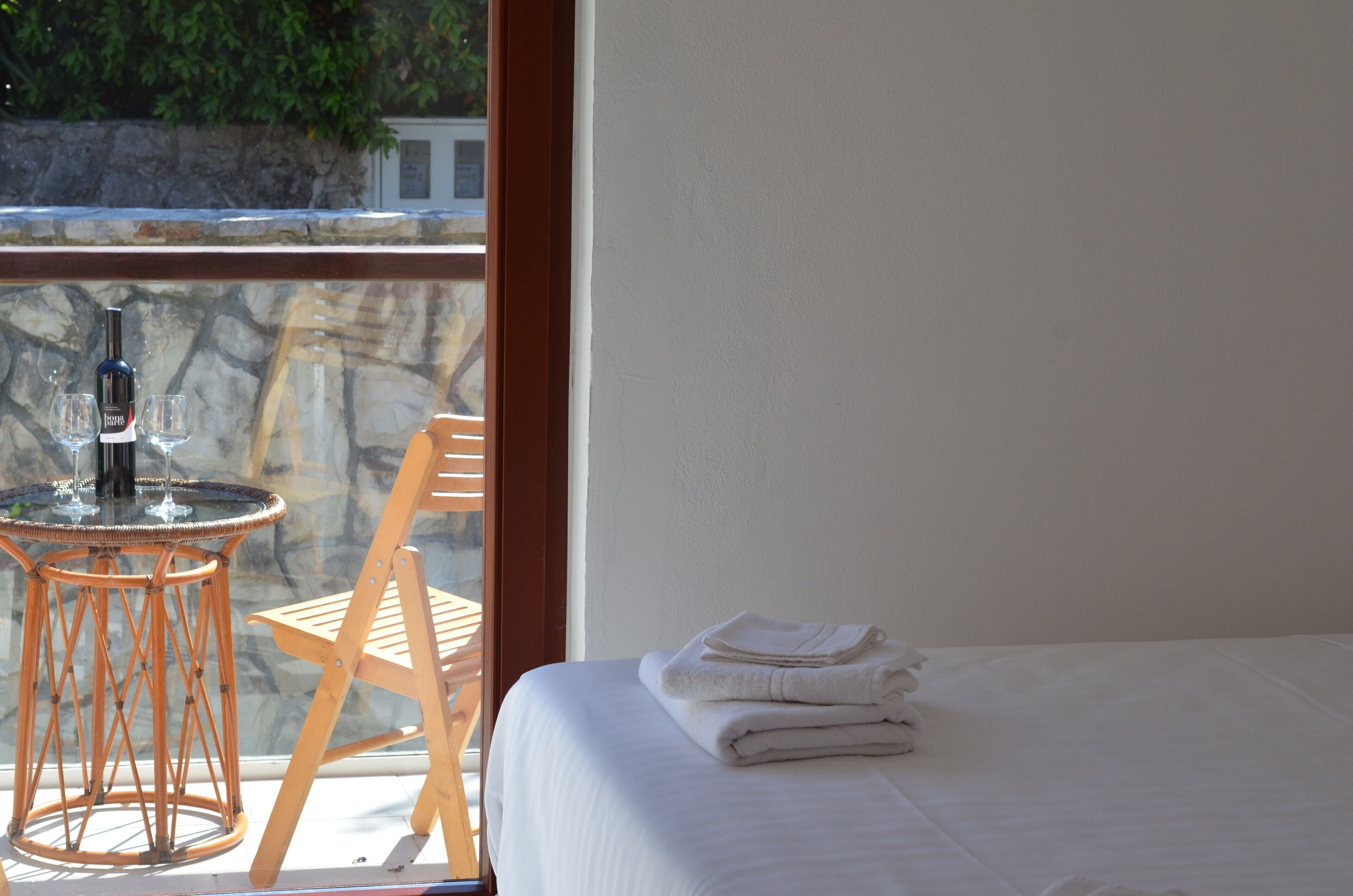 Habitación de hotel con una cama, una mesa con una botella y vasos, y vistas a un muro de piedra y follaje exterior.