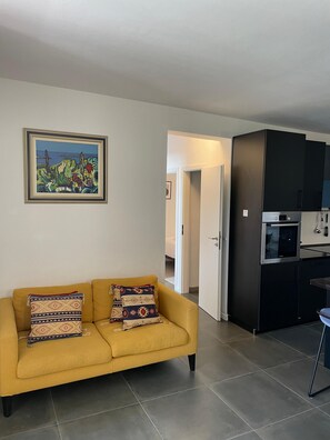 A living room with a sofa, a framed painting, and a kitchen with dark cabinets and appliances.