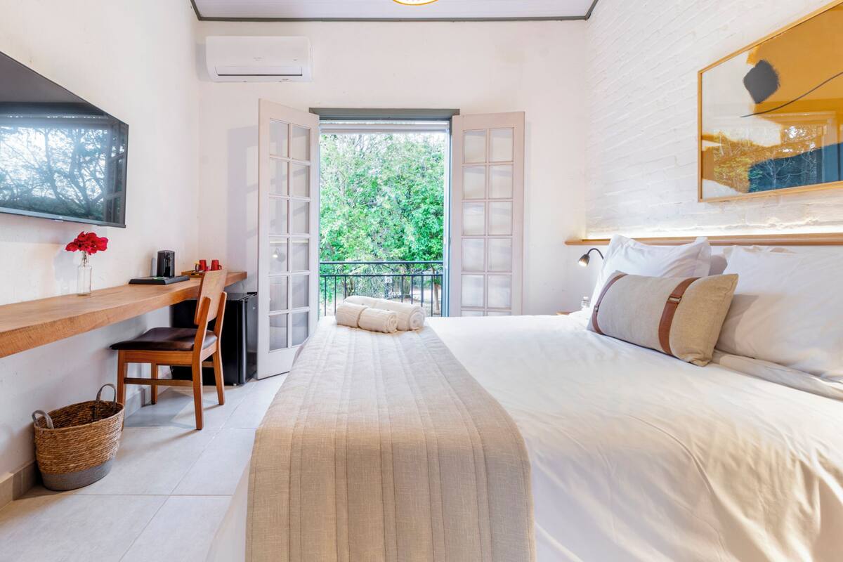 A bedroom with a bed, a TV, a desk, a chair, a basket, and a balcony with a view of trees.