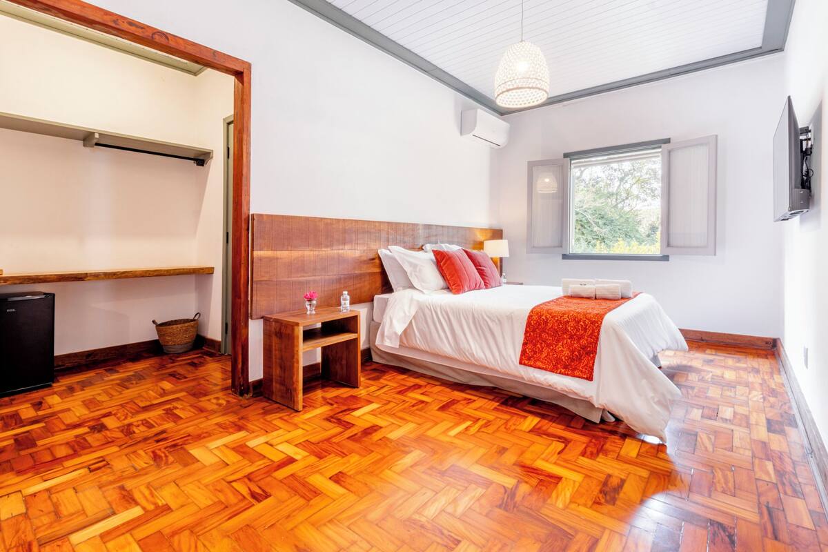 A bedroom with a wooden floor, a bed with white and red bedding, a bedside table with a lamp, a TV mounted on the wall, and a window with a view of greenery.