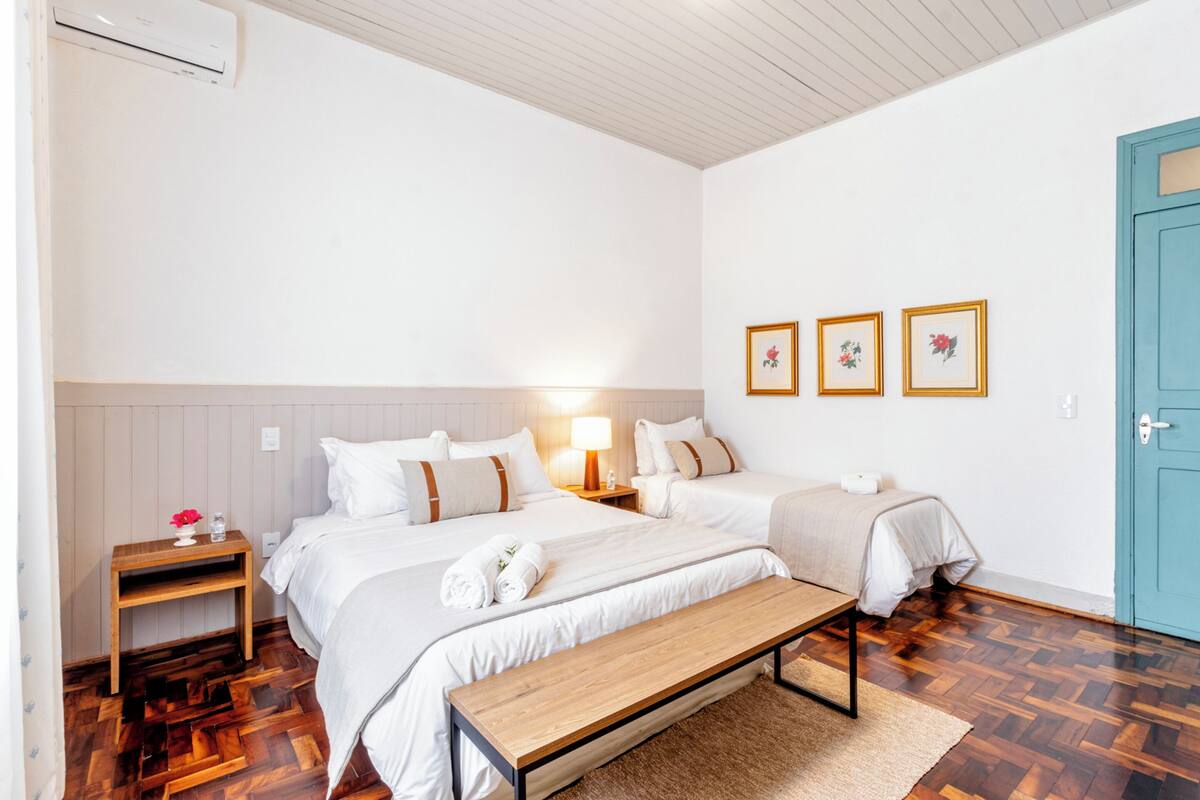 A hotel room with two beds, a wooden bench, a nightstand with a lamp, and a door with a blue handle.