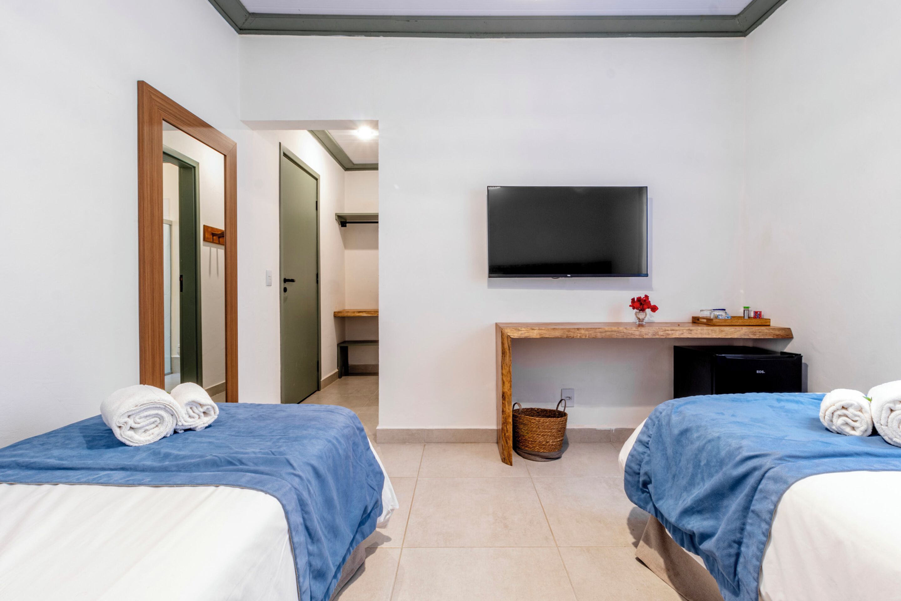 A hotel room with two beds, a TV mounted on the wall, a desk with a basket, and a door leading to another room.