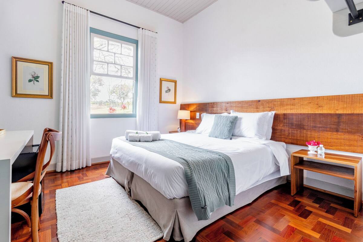 A neatly arranged bedroom with a wooden headboard, a bed with white linens, a bedside table with a lamp, and a view through a window with curtains.