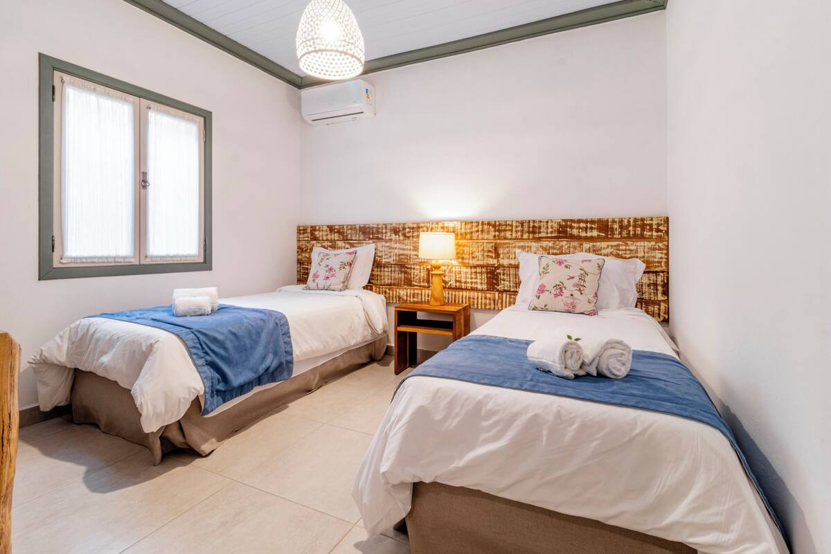 A hotel room with two beds, a window, an air conditioning unit, and a wall-mounted lamp.