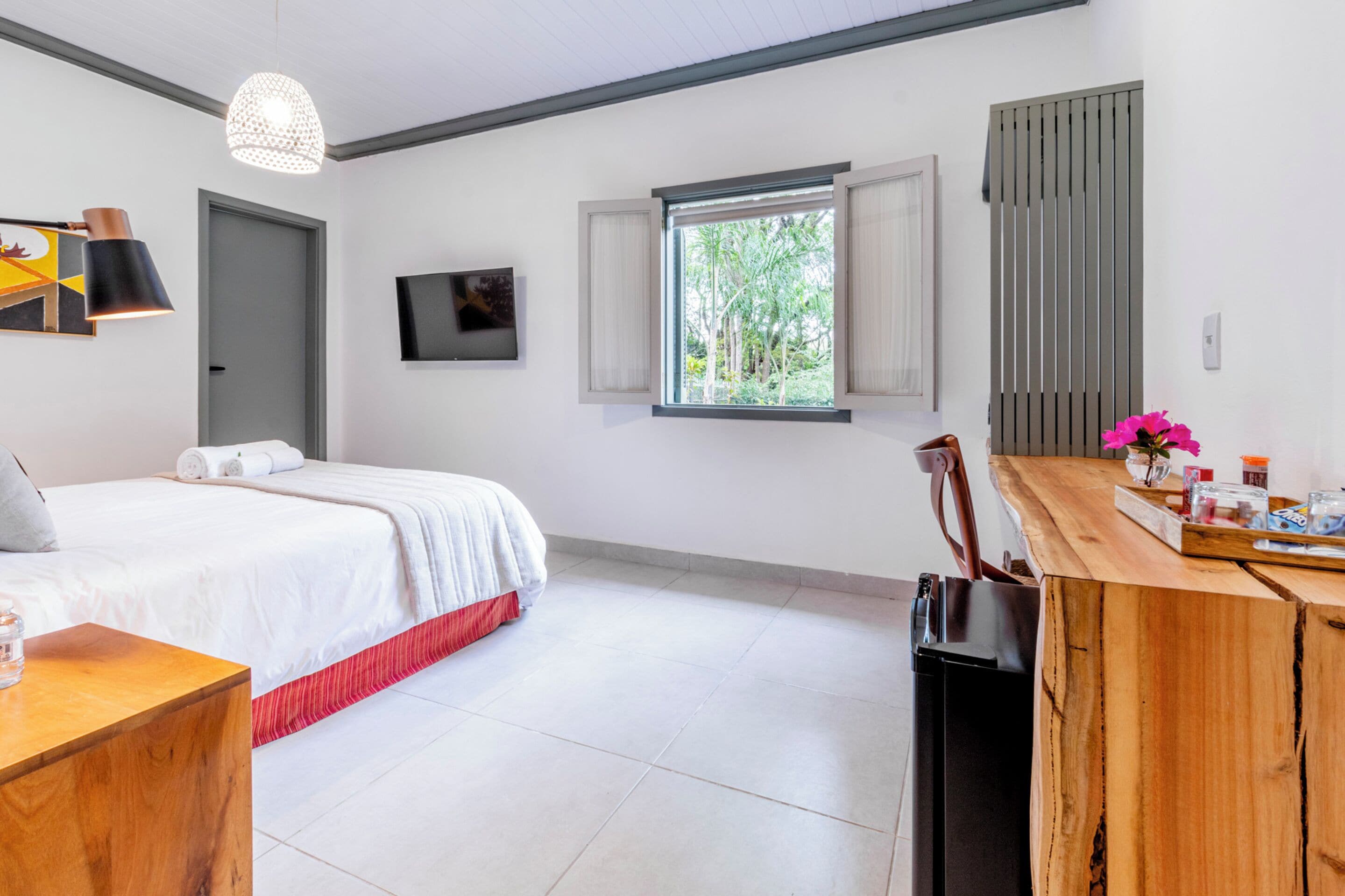 A modern bedroom with a bed, a desk, a TV, and a window with a view of greenery.
