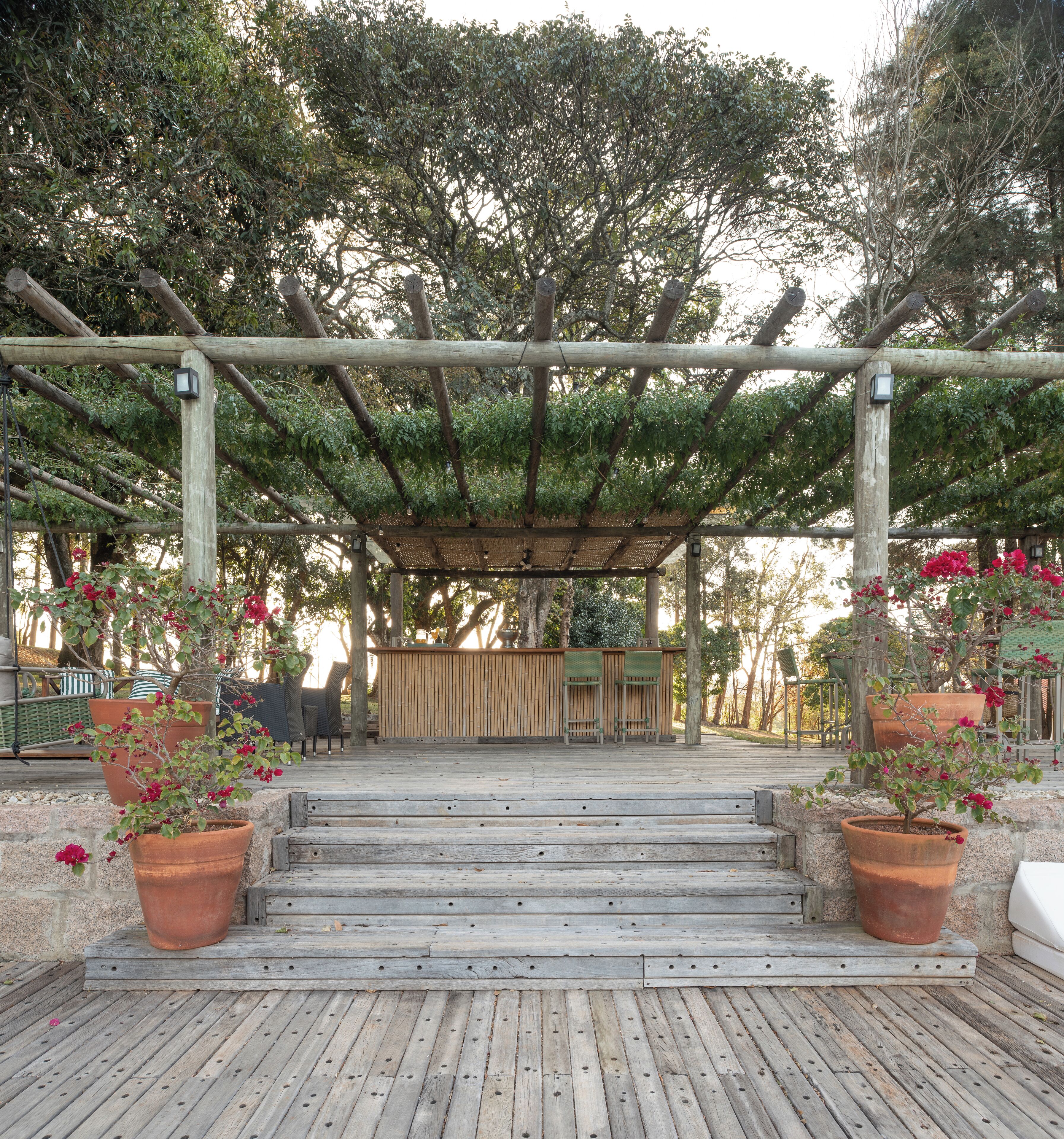 A wooden pergola with a seating area underneath, surrounded by potted plants and trees.
