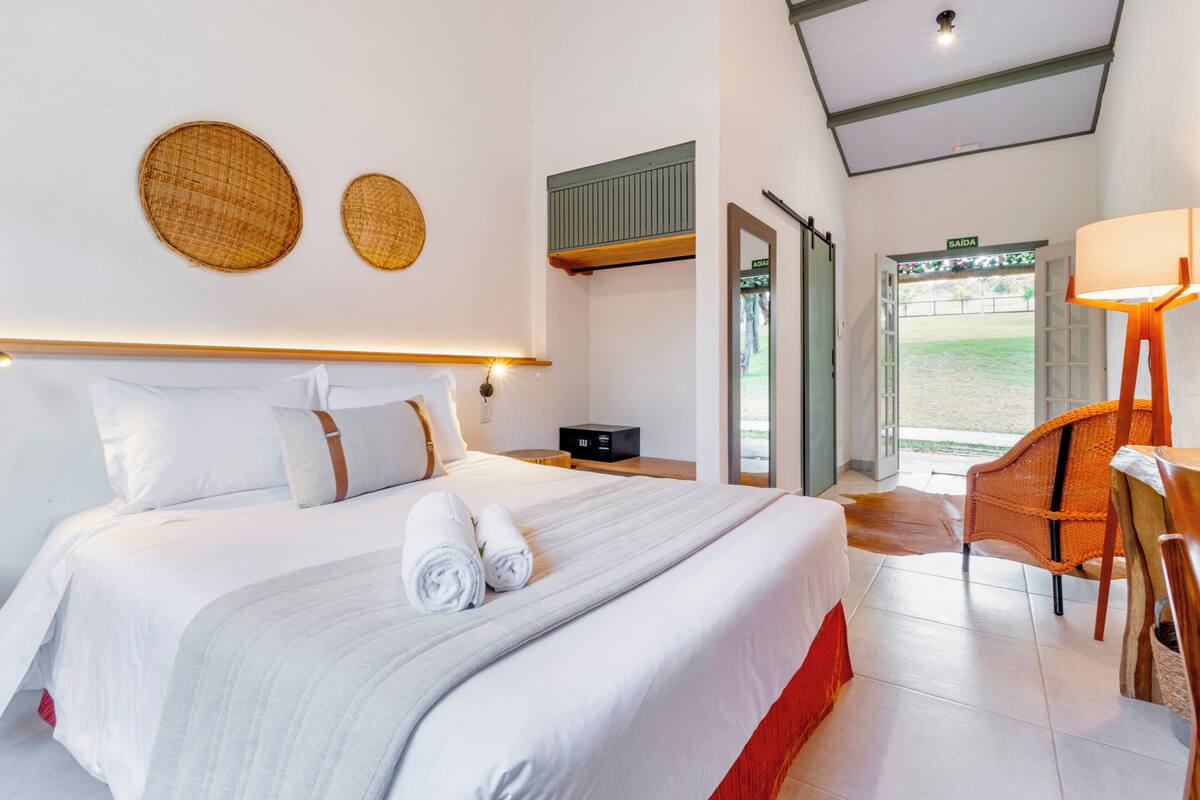 A hotel room with a large bed, a wooden headboard, a wicker wall decoration, a small table with a lamp, and a view of the outside through a door.