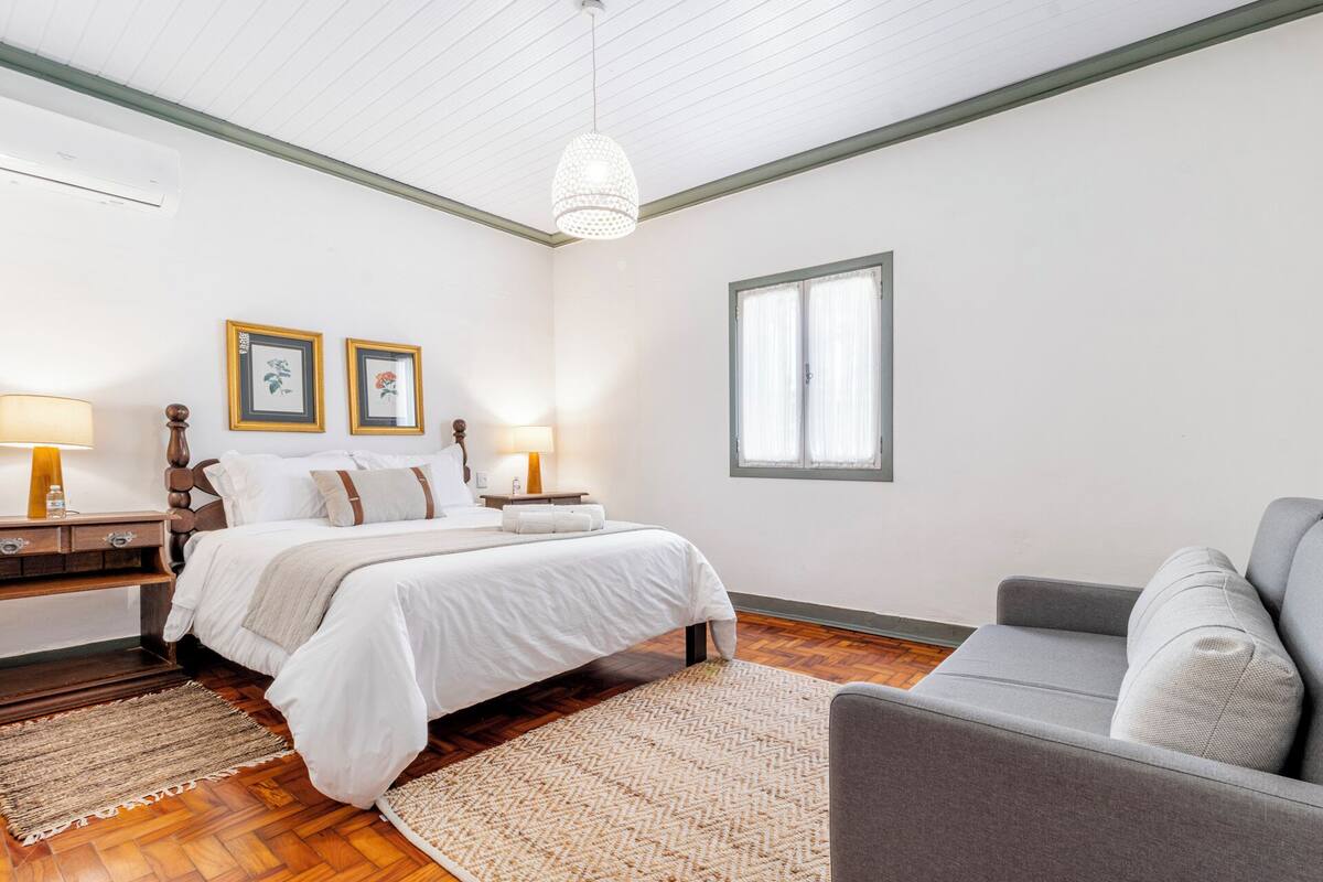 A bedroom with a bed, bedside tables, a grey armchair, a window, and a hanging light fixture.