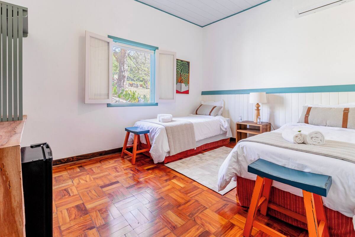 A hotel room with two beds, a wooden floor, a window with a view of greenery, and a small stool.