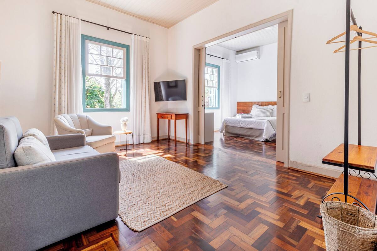 A spacious living area with a sofa, a television, a wooden table, and a bed in the background.