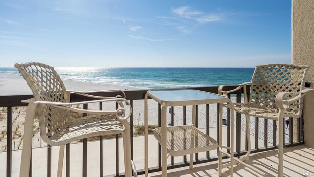A balcony with two wicker chairs and a small table, overlooking a beach and the ocean.