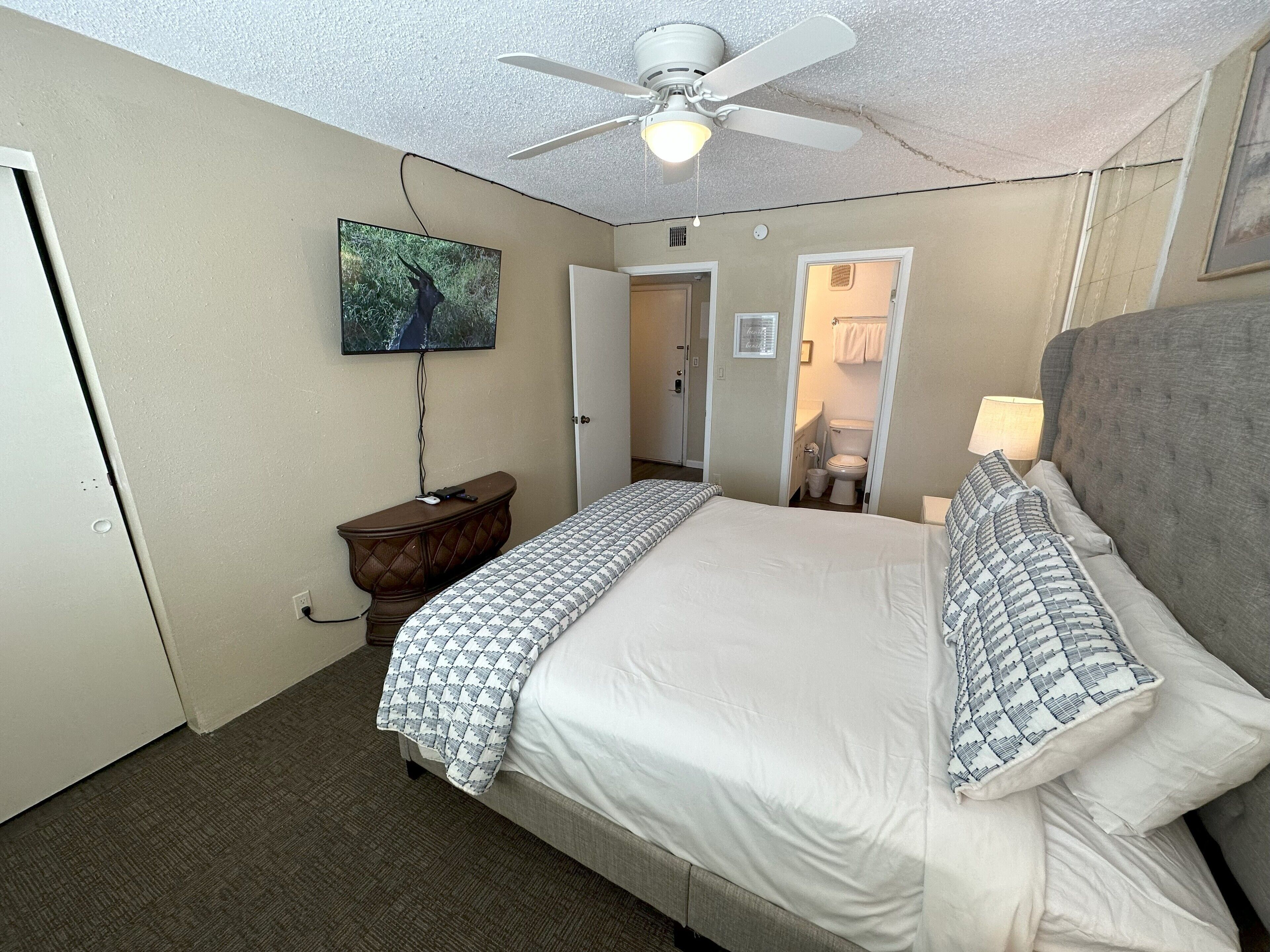 A hotel room with a large bed, a ceiling fan, a TV mounted on the wall, and a bathroom visible through an open door.