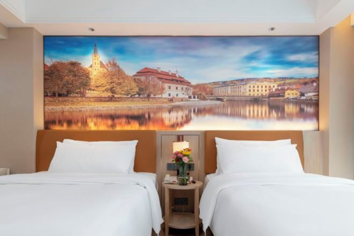 A hotel room with two beds, a large painting of a lake and buildings, and a small table with a vase of flowers.