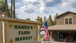 Uma placa de madeira para a Hanna's Farm Market com o logotipo de um girassol, uma bandeira americana e um edifício com uma placa que diz 'Farmers Market'.