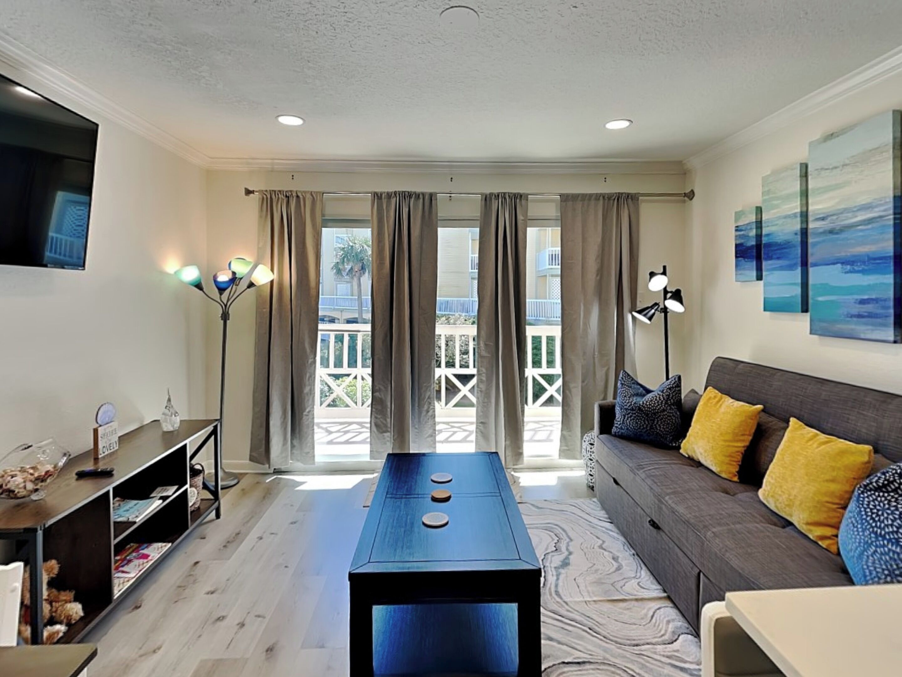 A modern living room with a sofa, a coffee table, and a TV. There are curtains, a floor lamp, and artwork on the wall.