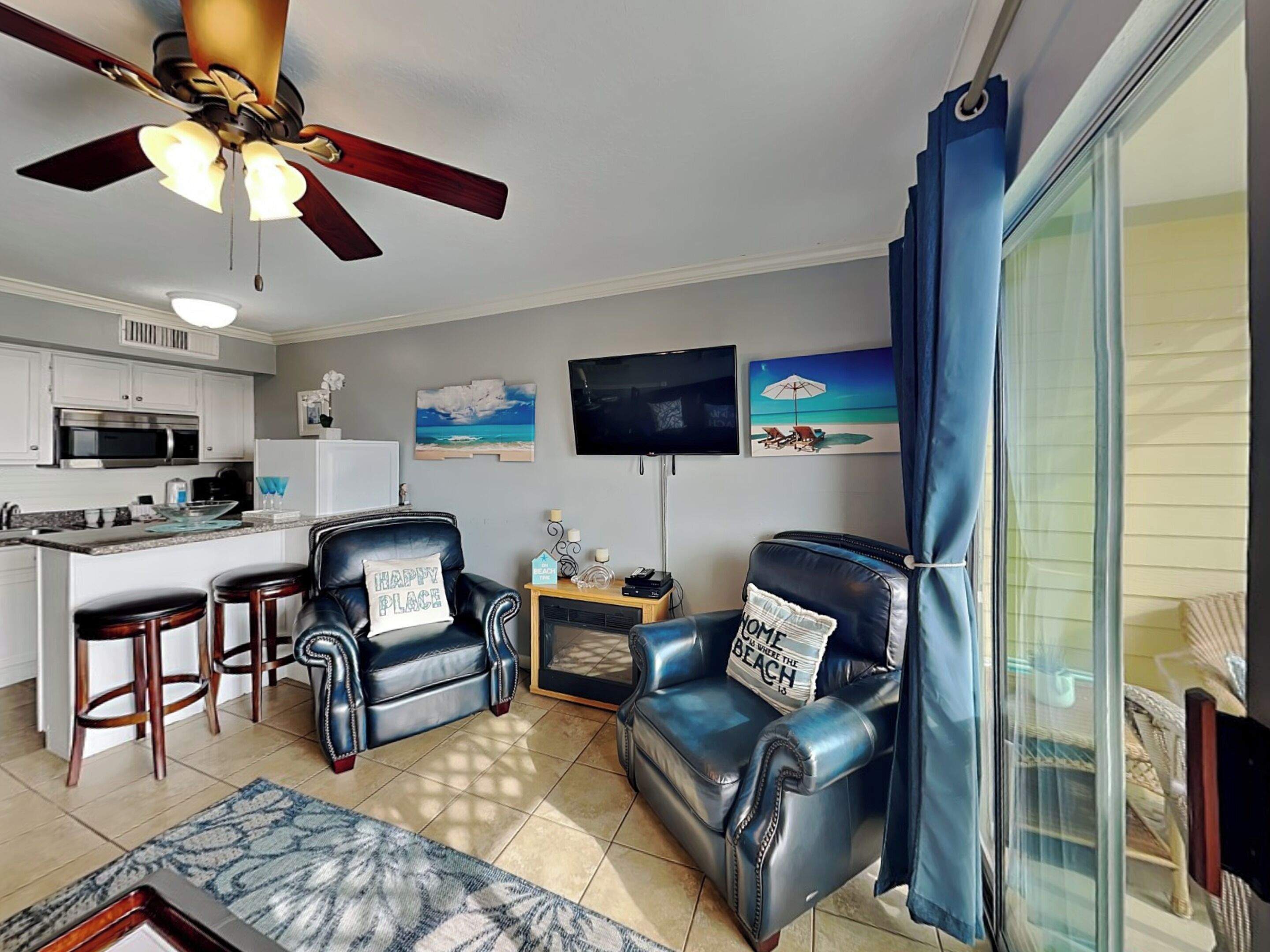 A living room with a kitchen, a ceiling fan, a TV, and a beach-themed painting.