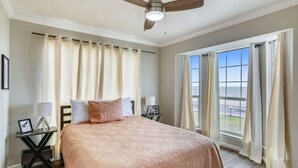 A bedroom with a bed, a ceiling fan, a nightstand, a window with curtains, and a view of the outside. - The Victorian by Ryson Vacation Rentals (Galveston)
