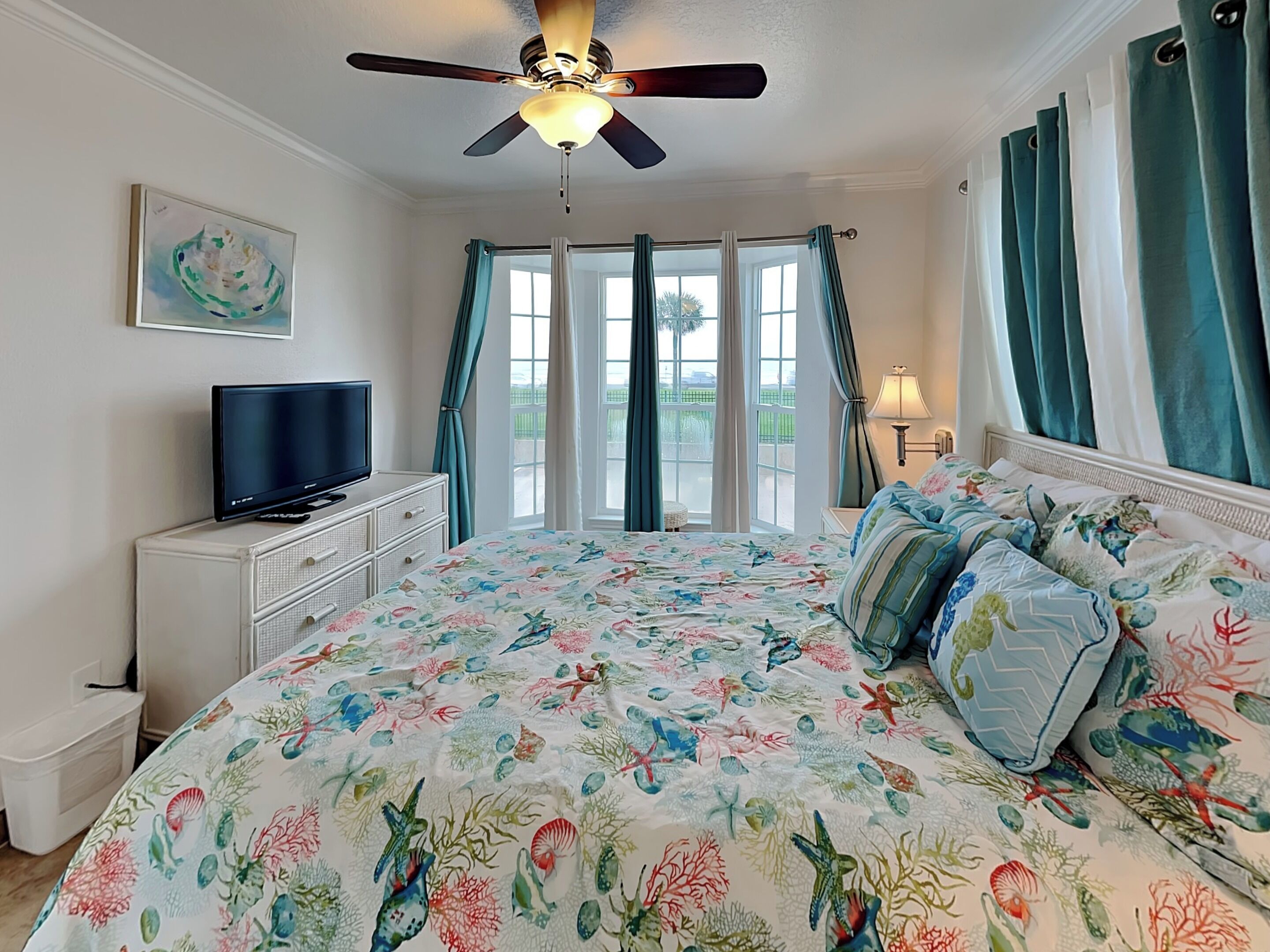 A bedroom with a bed, a television on a dresser, a ceiling fan, and a window with curtains.