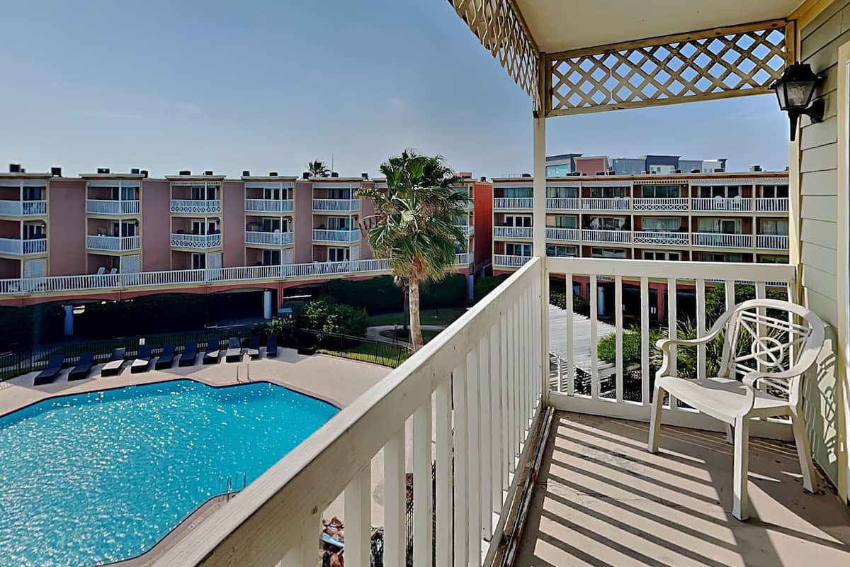 A balcony with a pool, white chairs, and a view of a multi-story building with balconies.