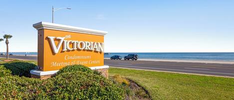 A sign for The Victorian Condominiums with Meeting & Event Centers, located near a road with vehicles and a beach in the background.