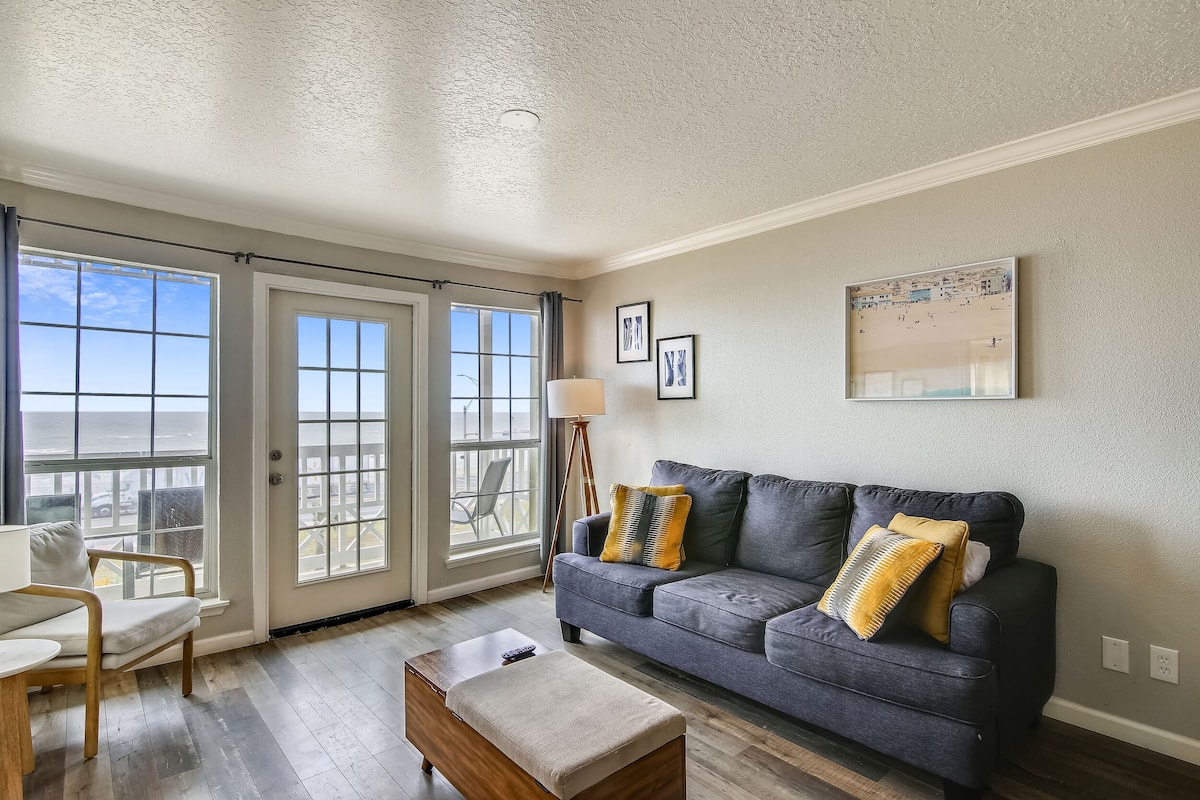 A living room with a gray sofa, a wooden coffee table, and a chair. There are two windows with curtains, a floor lamp, and framed pictures on the wall.