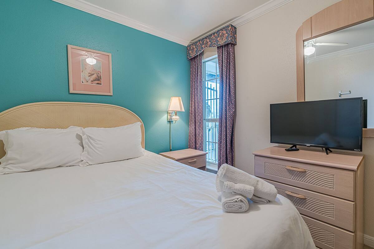 A neatly made bed with white linens, a television on a dresser, and a window with patterned curtains.