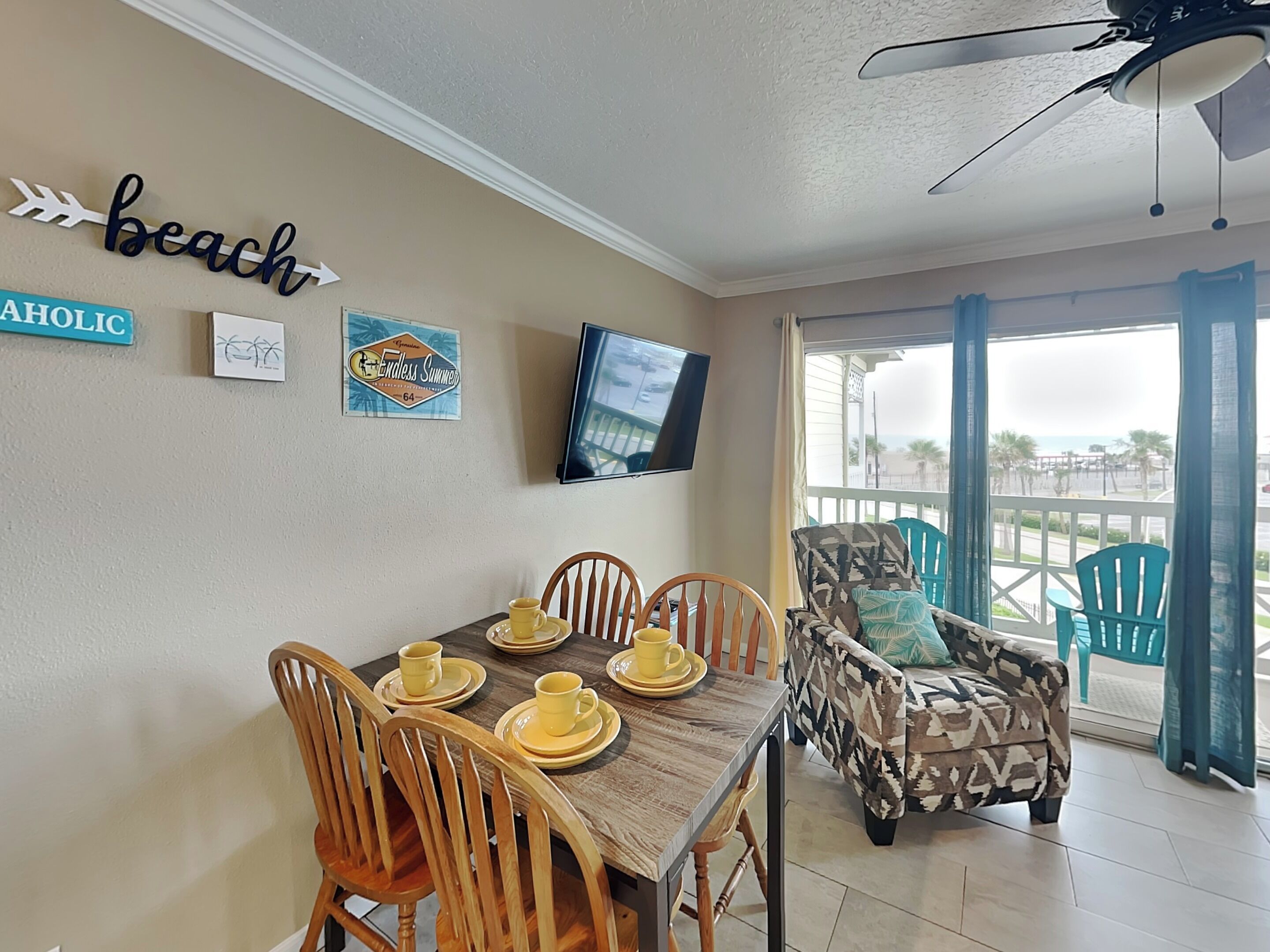 Un comedor con una mesa de madera dispuesta para cuatro personas, un televisor montado en la pared y vista a la playa a través de un ventanal grande.