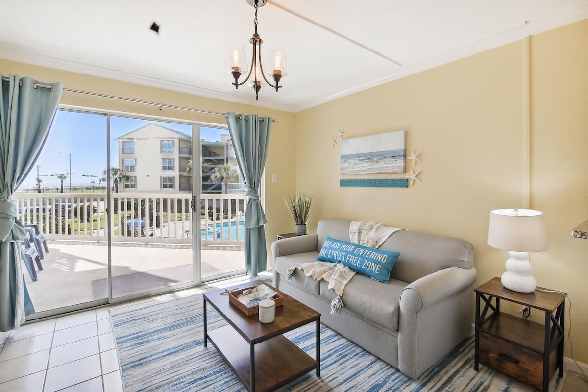 A living room with a sofa, a coffee table, a lamp, and a view of a pool and buildings outside.