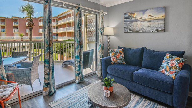 A living room with a blue sofa, a round coffee table with a plant, and a view of a balcony with outdoor furniture. - The Victorian by Ryson Vacation Rentals (Galveston)