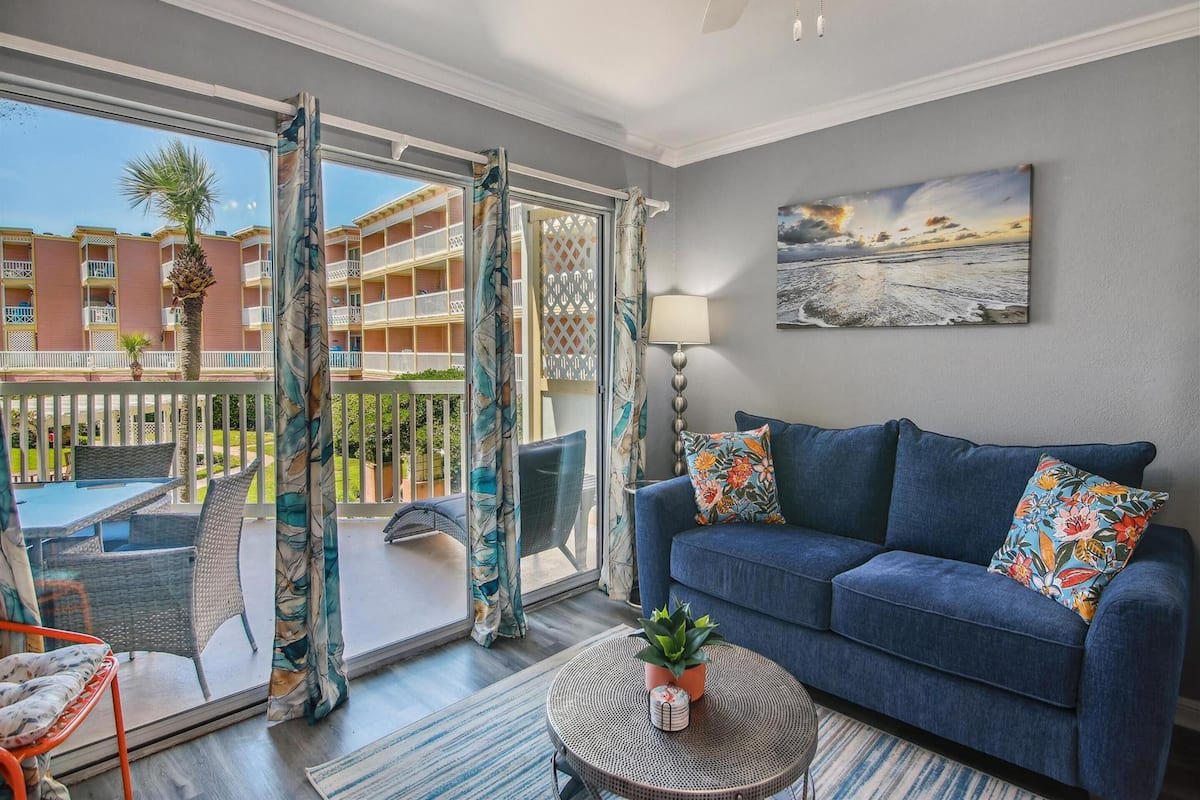 A living room with a blue sofa, a round coffee table with a plant, and a view of a balcony with outdoor furniture.
