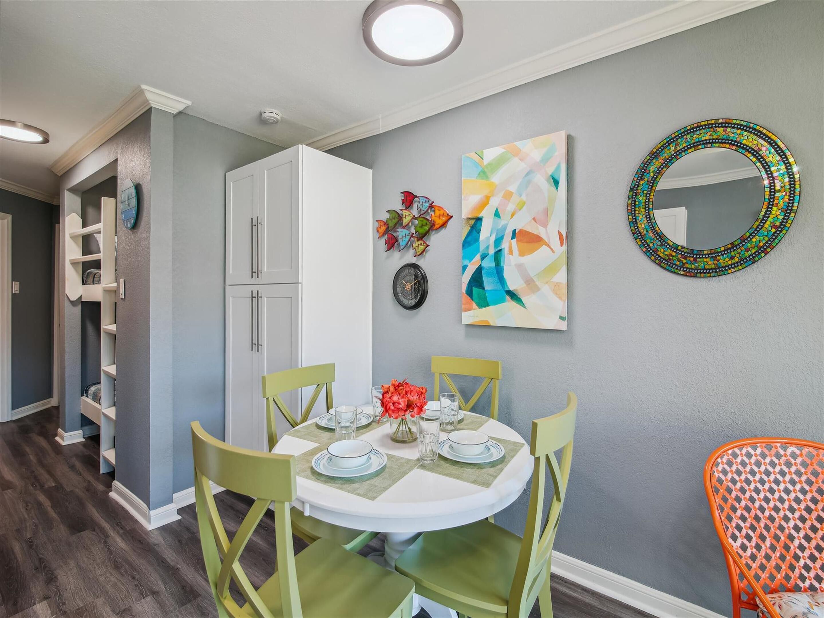 A dining area with a round table set for four, surrounded by green chairs. A white cabinet is visible to the left, and a colorful abstract painting hangs on the wall.