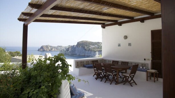 A covered outdoor seating area with a table and chairs, overlooking a coastal landscape with cliffs and the sea. - Domes, flowers and a small, cool pool (Le Forna)