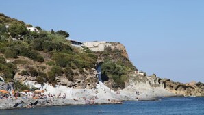A coastal area with rocky terrain, a cliff with buildings, and a beach with people and umbrellas. - Domes, flowers and a small, cool pool (Le Forna)