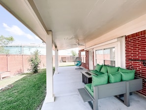 A covered patio with a grey sofa and cushions, a hanging chair, and a red brick wall.