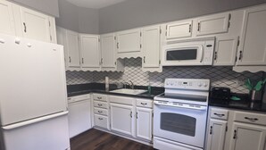 Une cuisine avec des meubles blancs, un plan de travail noir, un réfrigérateur blanc, un four à micro-ondes, une cuisinière et un évier.