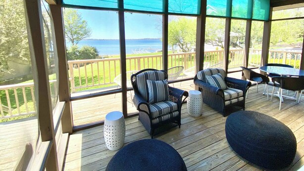 Een overdekt terras met rieten stoelen en poefs, met uitzicht op een houten terras en het water.