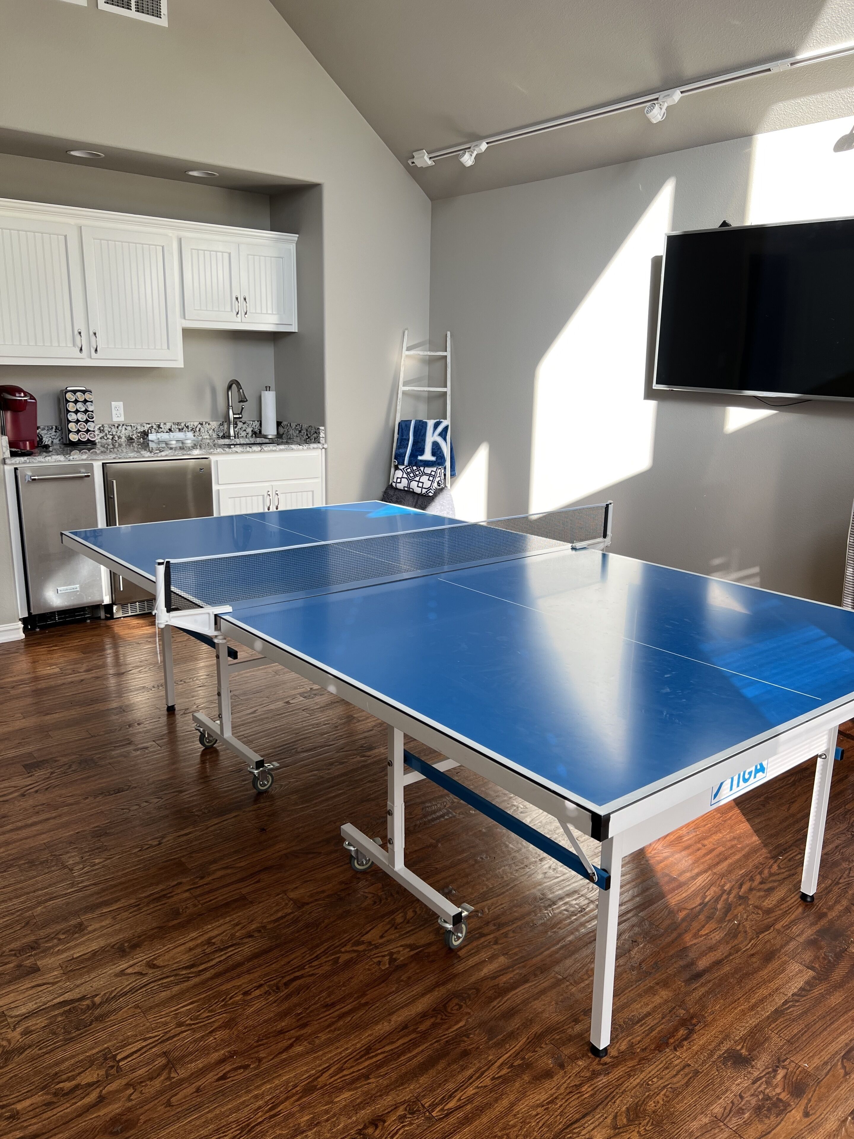 A ping pong table in a room with wooden flooring, white cabinets, a kitchen area, and a mounted TV.