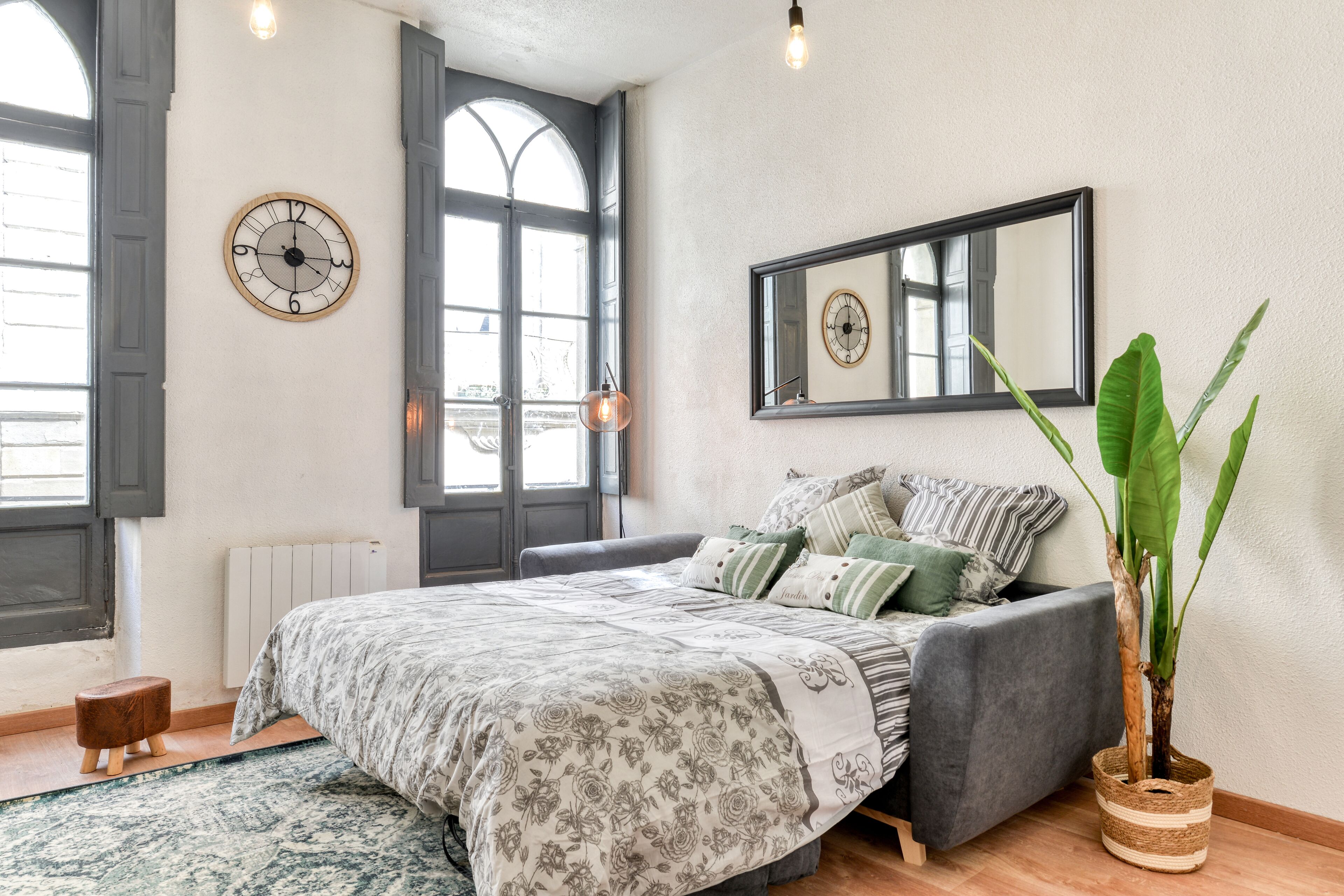 Un dormitorio con cama, un ventanal grande, un reloj de pared, un espejo y una planta en maceta.