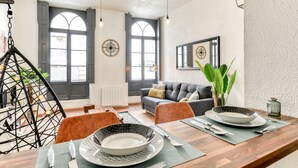 Ein modernes Wohnzimmer mit Essbereich, ausgestattet mit einer Couch, Sesseln, einer Hängeleuchte und einem großen Fenster mit Fensterläden.