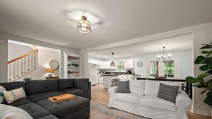 A spacious living area with a dark grey sectional sofa, a white sofa, and a wooden coffee table. A kitchen with white cabinets and a central island is visible in the background.