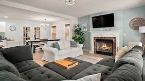 A modern living room with a fireplace, a flat-screen TV, and a dining area in the background.