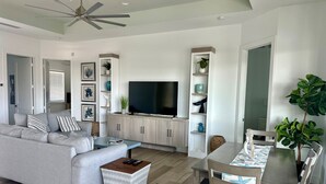 Una sala de estar moderna con un sofá gris, un televisor sobre un mueble de madera, una mesa de comedor con sillas y un ventilador de techo.