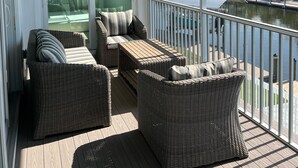 Un espacio al aire libre techado con mobiliario de mimbre, ventilador de techo y vistas a un cuerpo de agua y edificios.