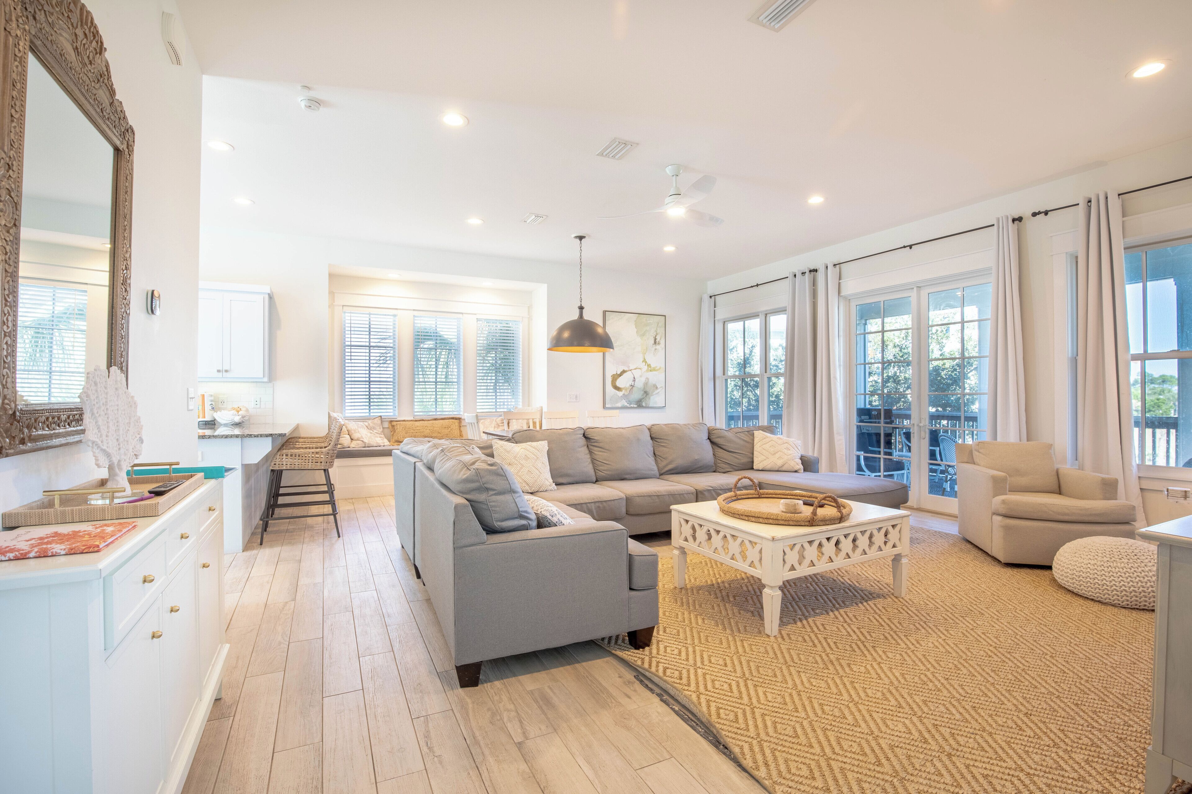 A spacious living room with a grey sofa, a white coffee table, and a large window with curtains.